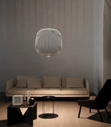 En minimalistisk stue med Spokes pendellampe fra Foscarini som et elegant midtpunkt. Lampens filigrane, metalliske stag kaster myke lysmønstre på veggen og taket, og gir rommet en lett, skulpturell effekt. Lys sofa, mørke aksenter og naturlige materialer skaper en rolig, moderne atmosfære