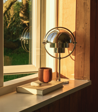 Forkrommet bordlampe i en vinduskarm i kveldslyset, ved siden av en bok og et keramikkrus.