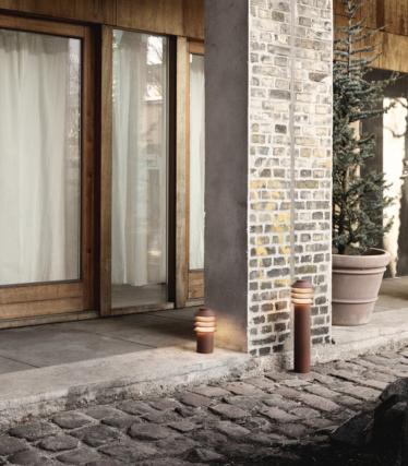 Uteområde med to moderne sylindriske gangbelysningsarmaturer plassert ved siden av en steinbelagt gangvei og en søyle av murstein og betong. Bak dem står glassdører med treramme, lysgardiner og en stor potteplante. Det varme lyset fra armaturene skaper en koselig og innbydende atmosfære.