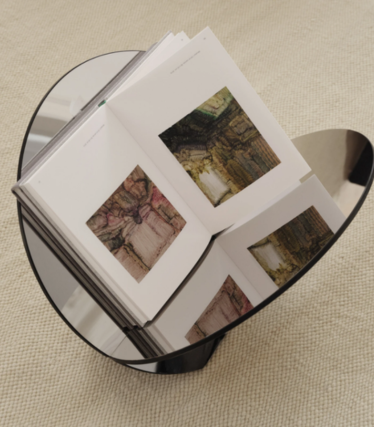 Rundt sidebord i mørkt glass med en åpen kunstbok, på et beige teppe.