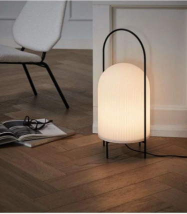 Hvit rillet lampe med svart bøylehåndtak på fiskebeinsparkett ved siden av en lenestol.